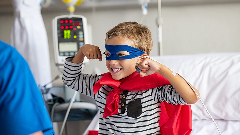 Strong young boy in hospital bed dressed as superhero.